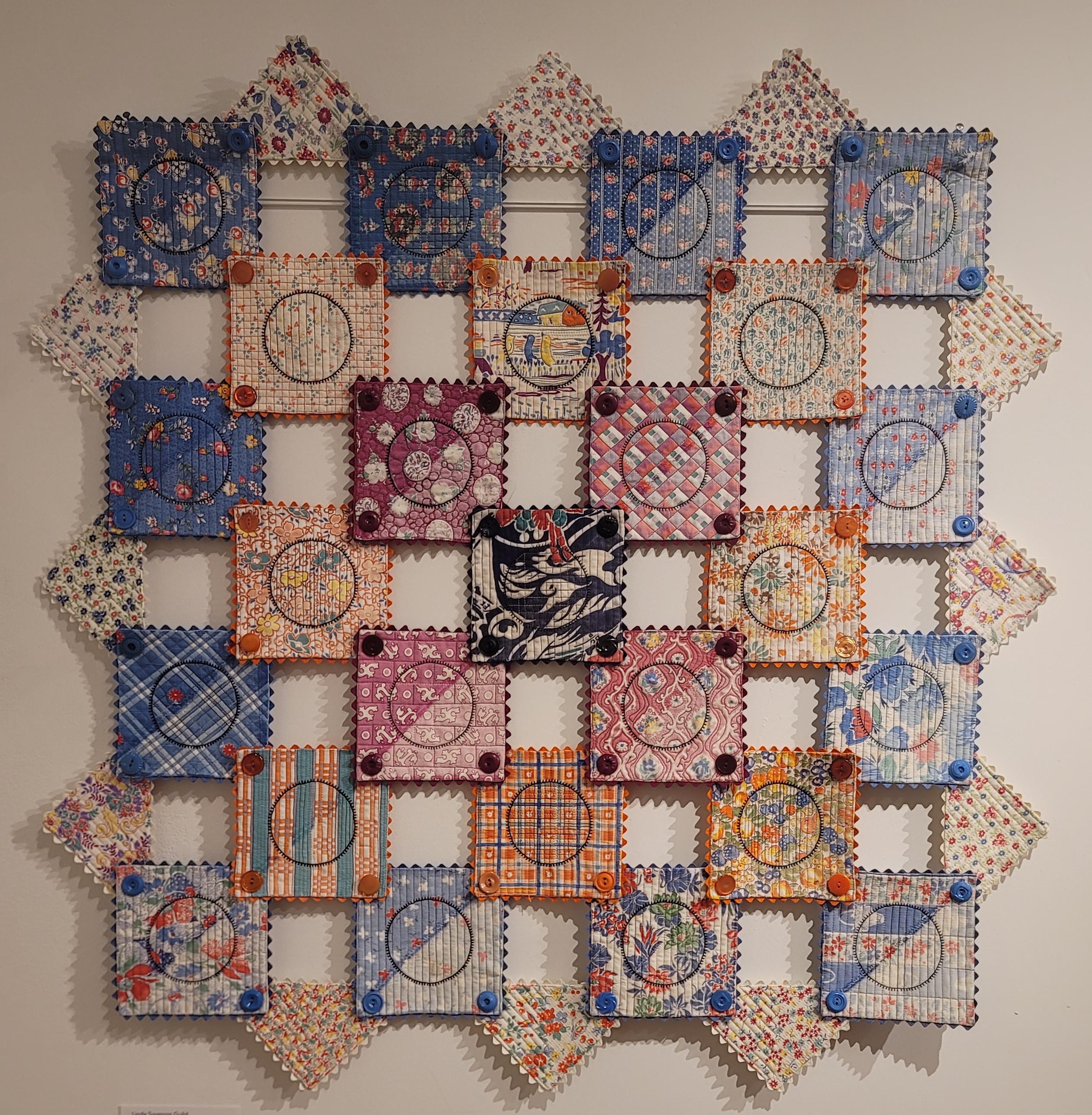 Refined Comfort by Linda Syverson Guild. A grid of mostly disconnected quilt squares attatched by buttons and a circle in the middle